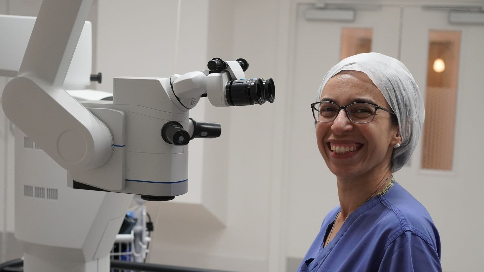 Ms. Nardine Menassa, Consultant Ophthalmic Surgeon, smiling in surgical scrubs and a cap beside an operating microscope in the theatre at Spire Liverpool Hospital.