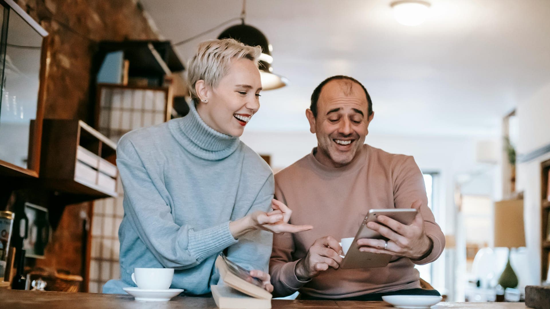 Two adults smile and interact while looking at a tablet at a cozy café setting.