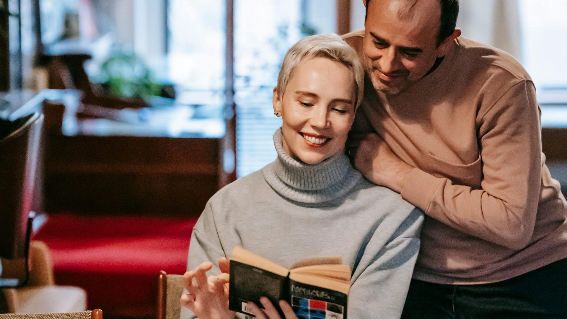A woman reads a book while a man embraces her from behind in a cozy indoor setting.