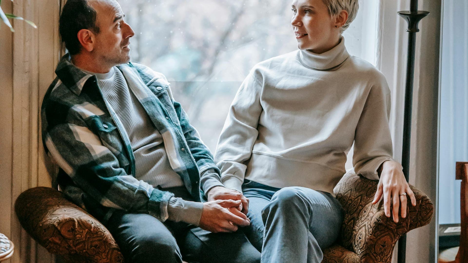 A man and a woman sit together on a sofa, holding hands and looking at each other, with a window in the background.