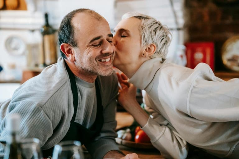 A woman kisses a smiling man while they are in a kitchen filled with food.