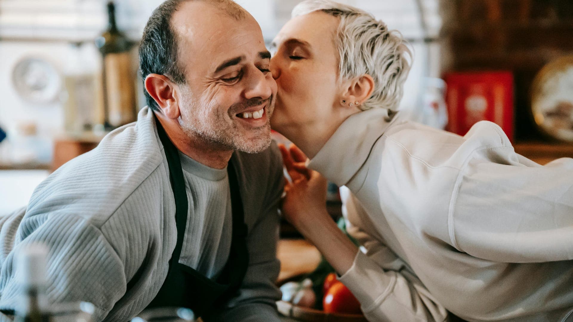 A woman kisses a smiling man while they are in a kitchen filled with food.