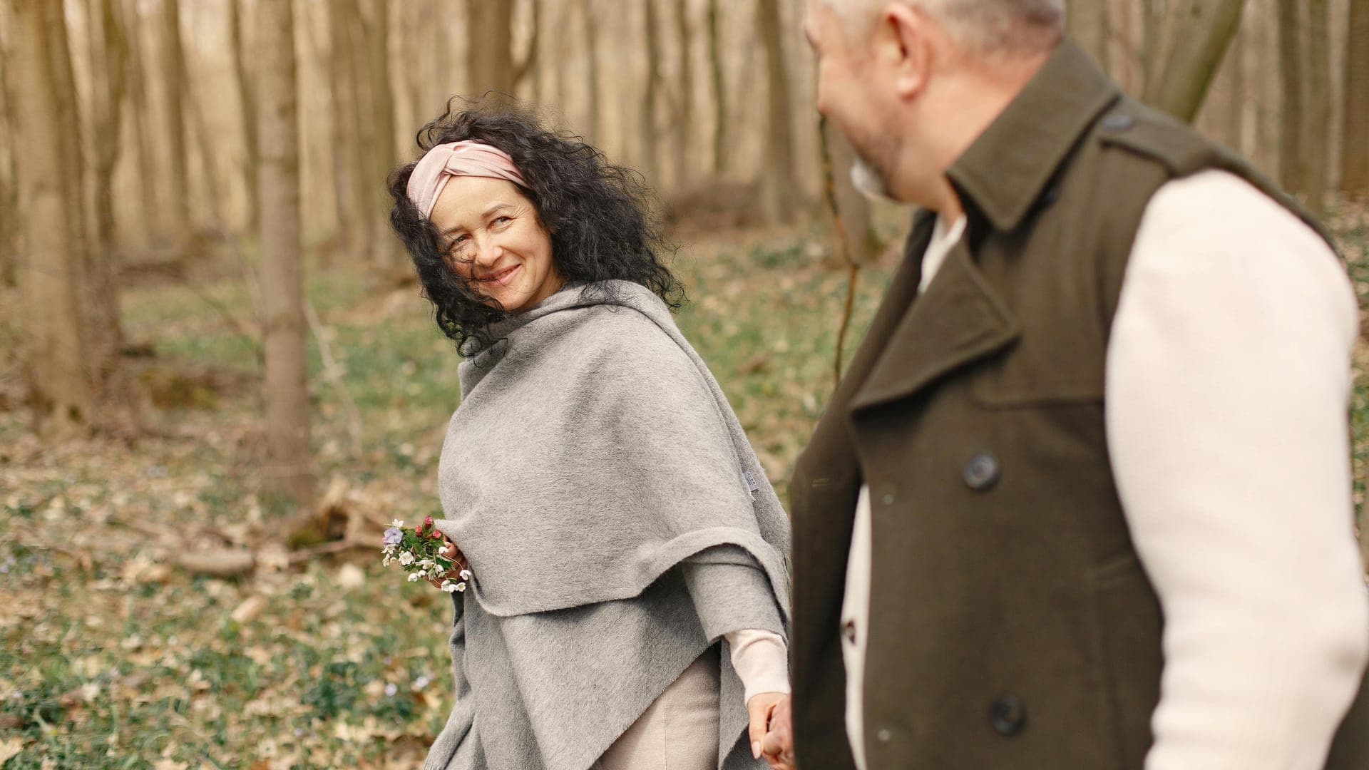 A woman in a gray poncho and a headband smiles at a man while walking in a forest.