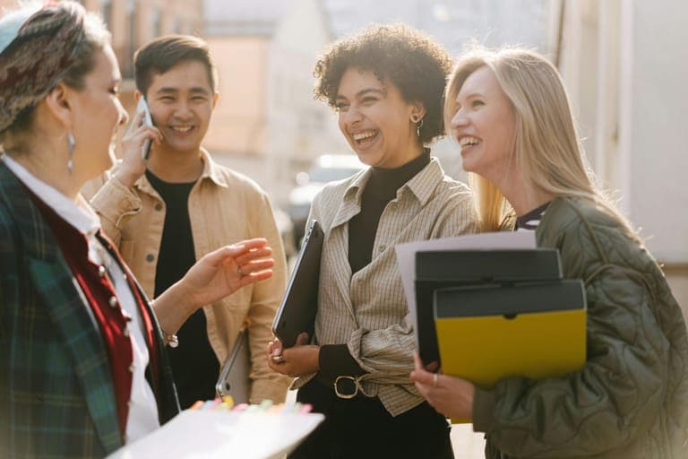 A group of four young adults laughing and talking together outdoors, holding folders and one person using a phone.