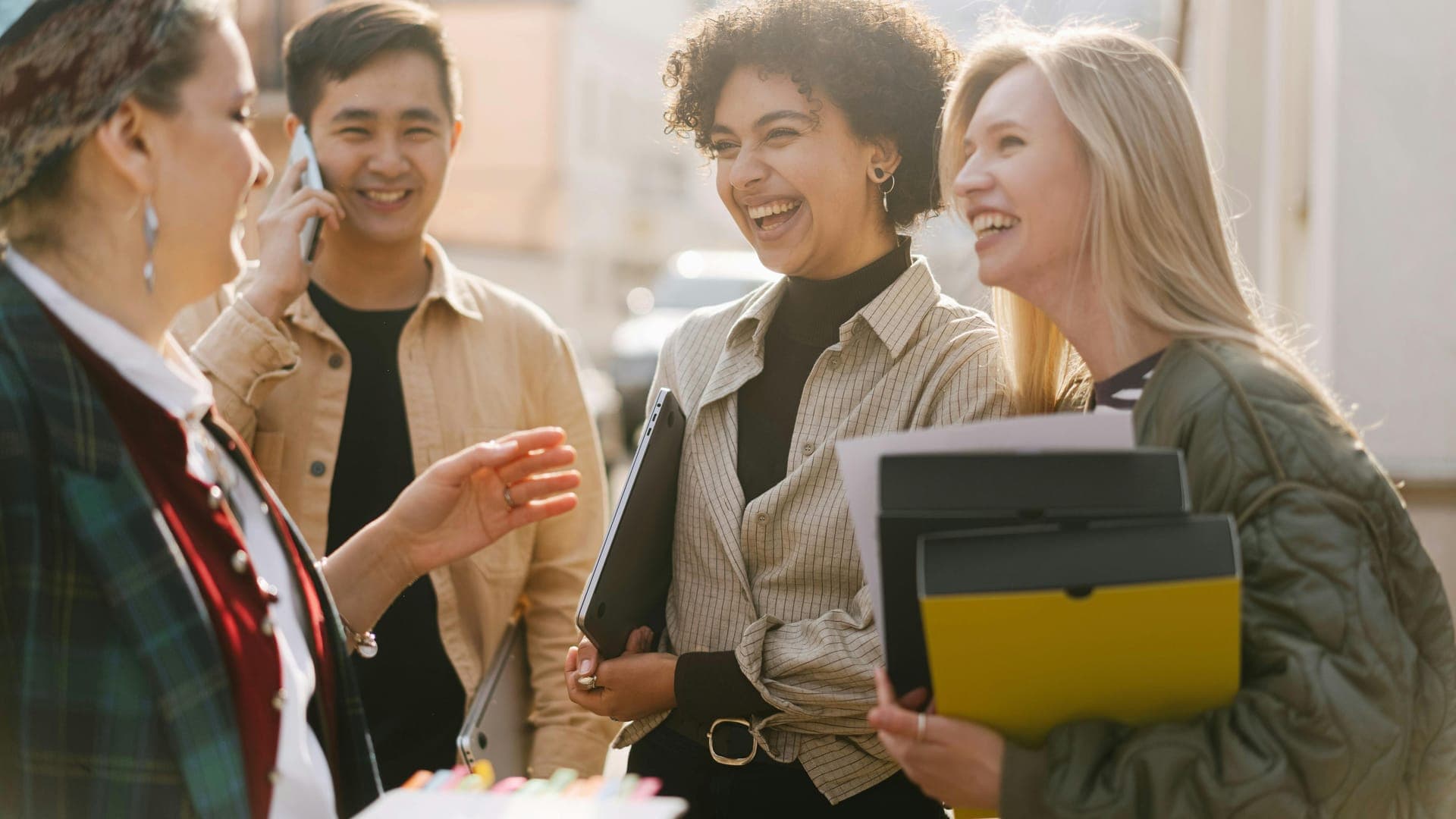 A group of four young adults laughing and talking together outdoors, holding folders and one person using a phone.