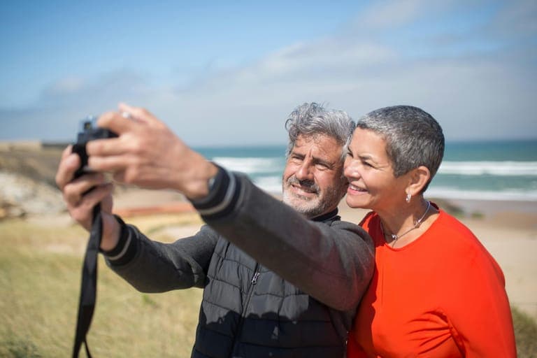 A smiling man and woman take a selfie together with a beach and ocean in the background.