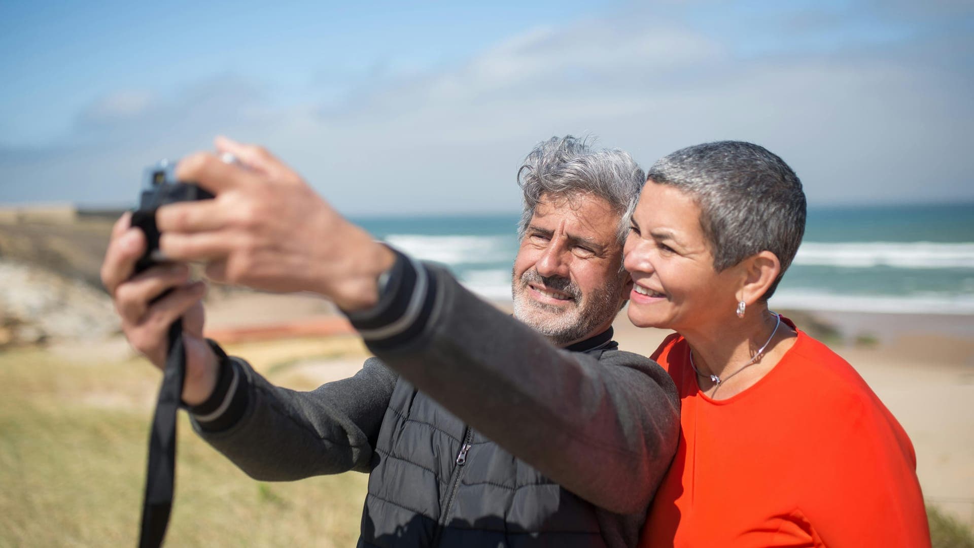 A smiling man and woman take a selfie together with a beach and ocean in the background.