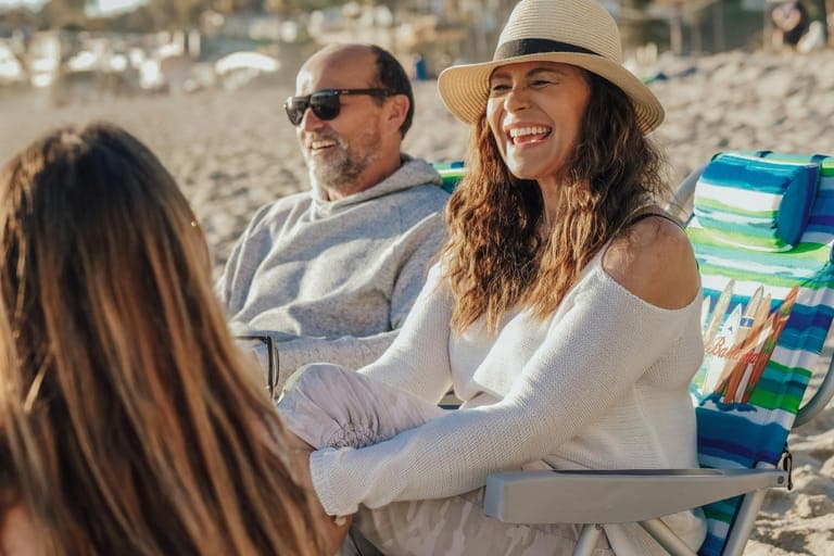 Two people sitting on a beach, smiling and enjoying the sun, with beach chairs and sand in the background.