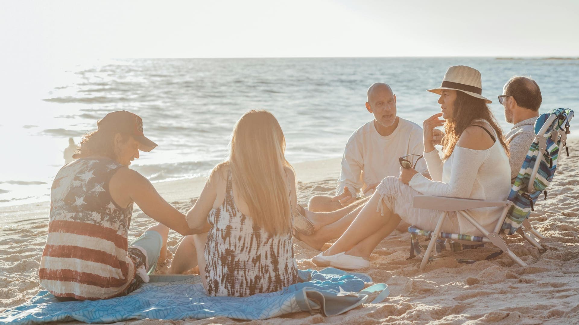 A group of five people sits on the beach, engaged in conversation with the ocean in the background.