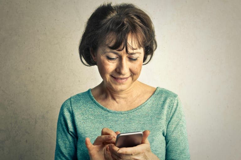 A smiling woman with shoulder-length hair interacts with a smartphone while wearing a light blue sweater.