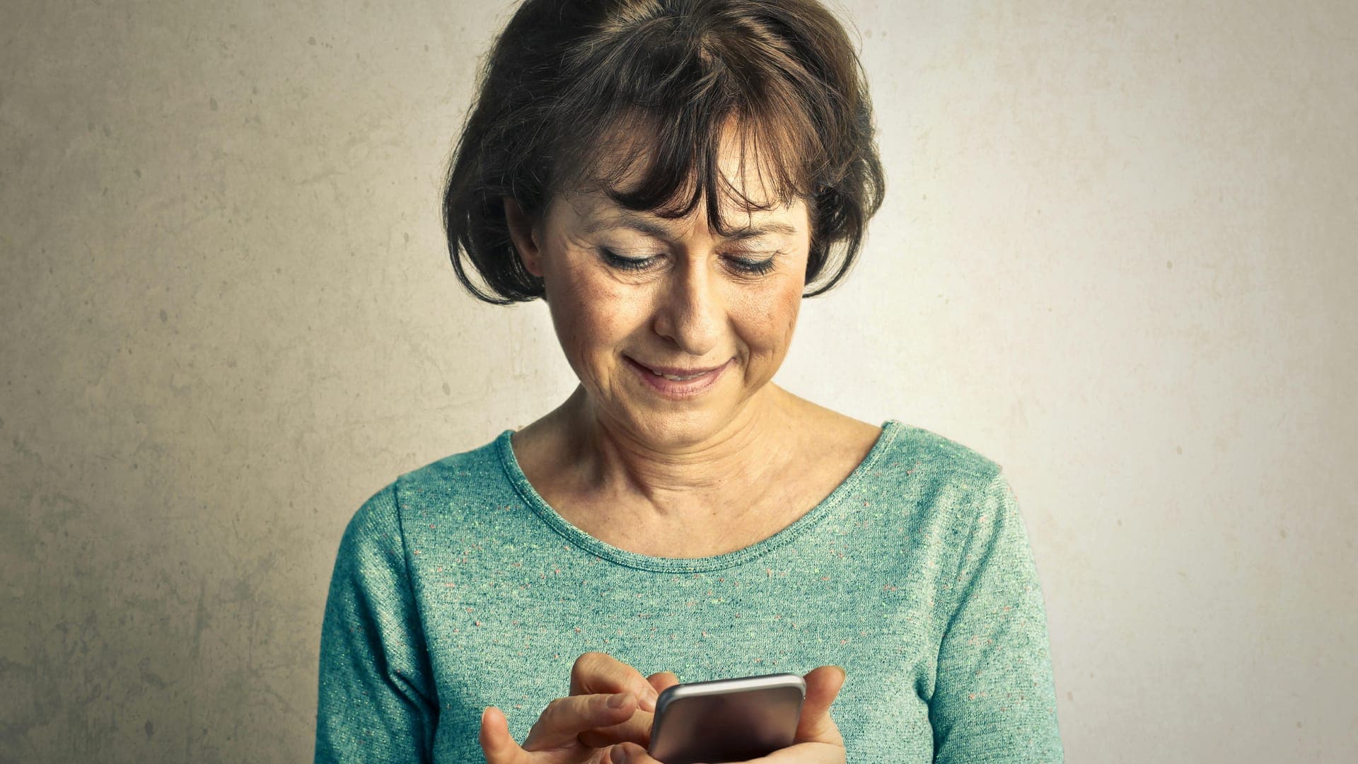 A smiling woman with shoulder-length hair interacts with a smartphone while wearing a light blue sweater.