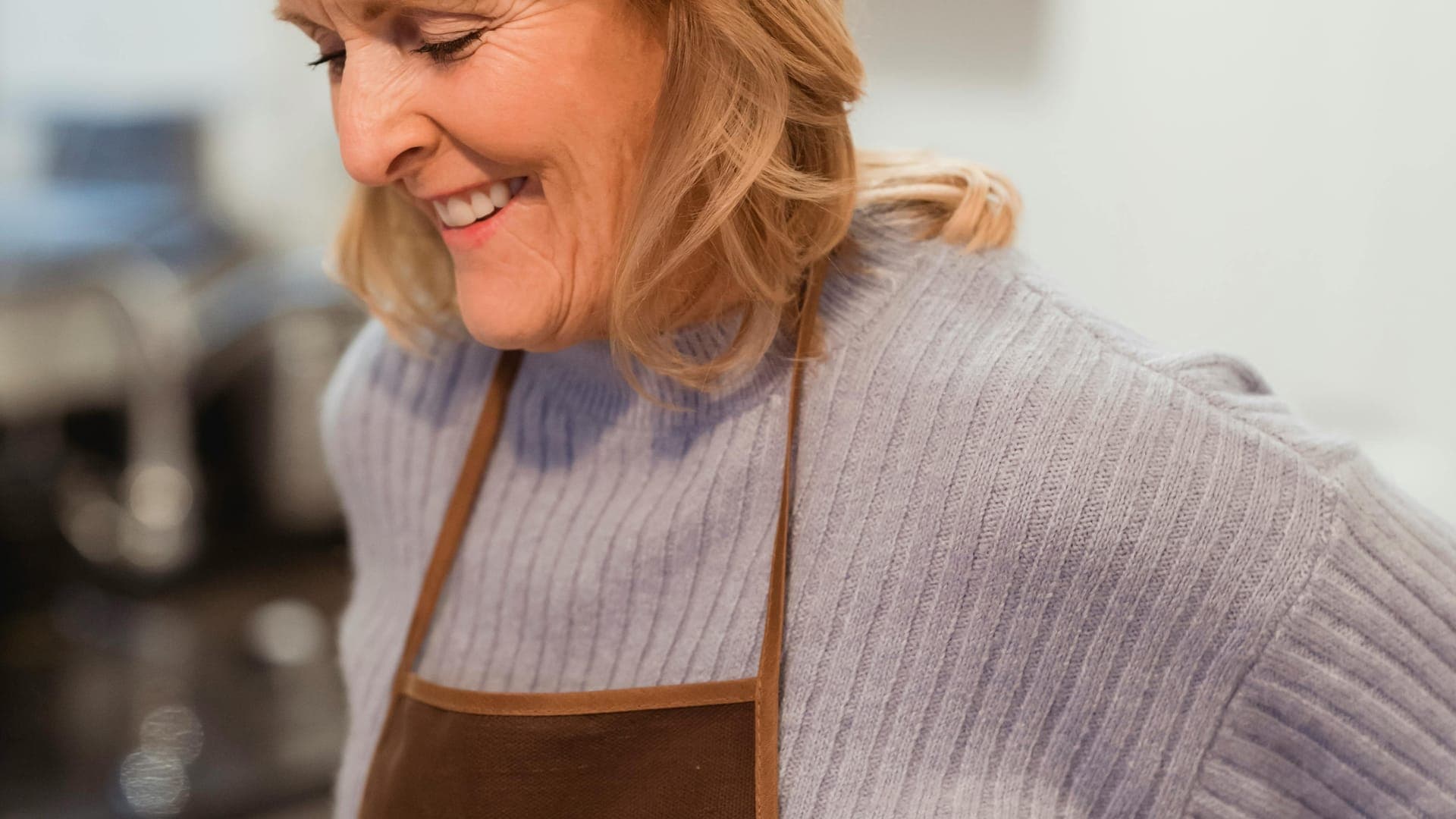 A smiling woman in a blue sweater and brown apron stands in a kitchen.