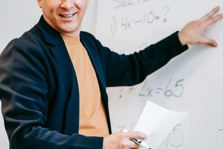 A smiling man points to a whiteboard with math equations while holding a piece of paper and a pen.
