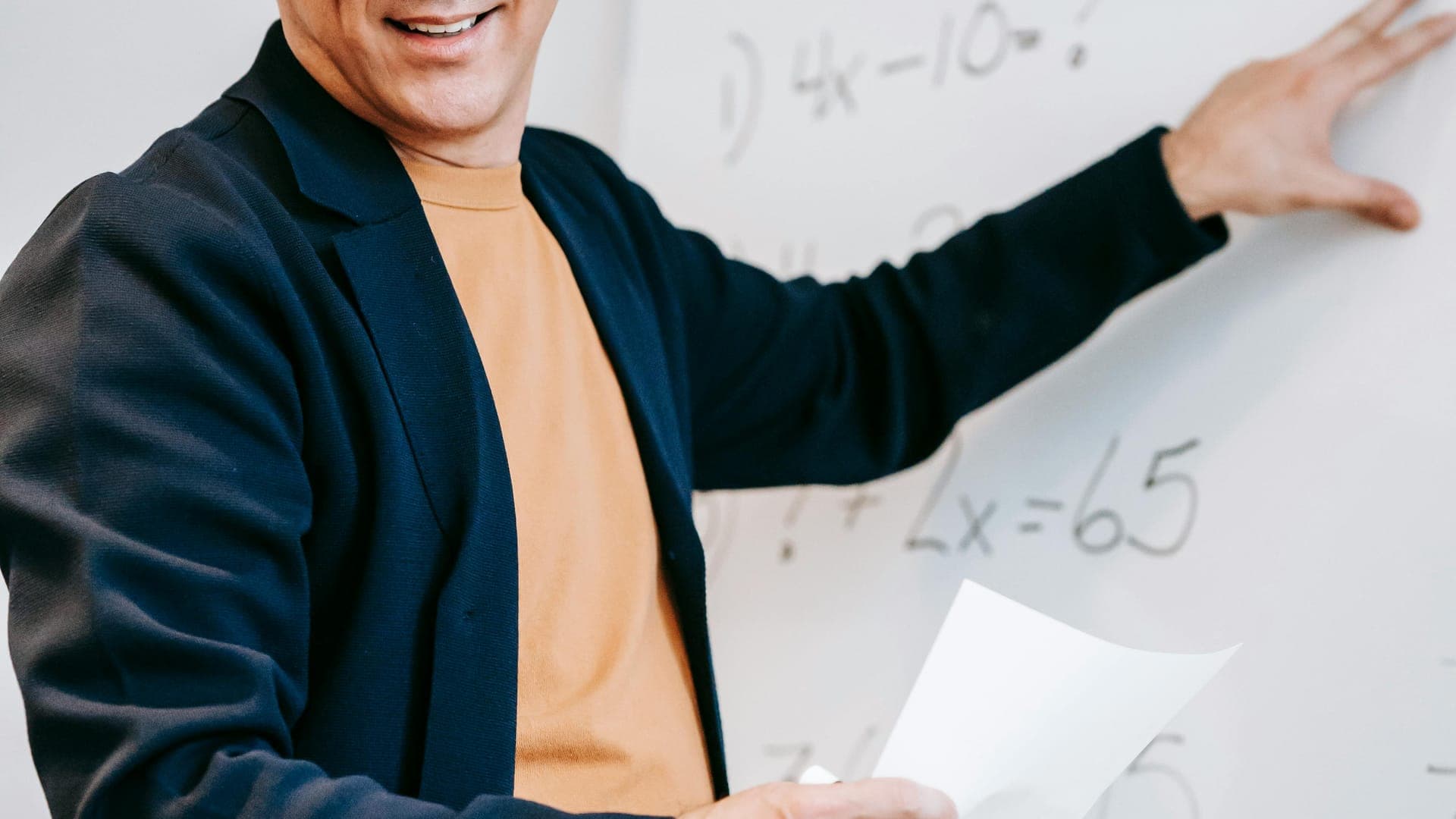 A smiling man points to a whiteboard with math equations while holding a piece of paper and a pen.