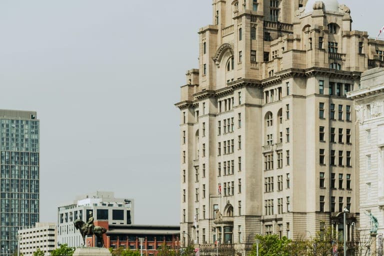 Historic clock tower building beside a paved walkway with people walking and sitting nearby.