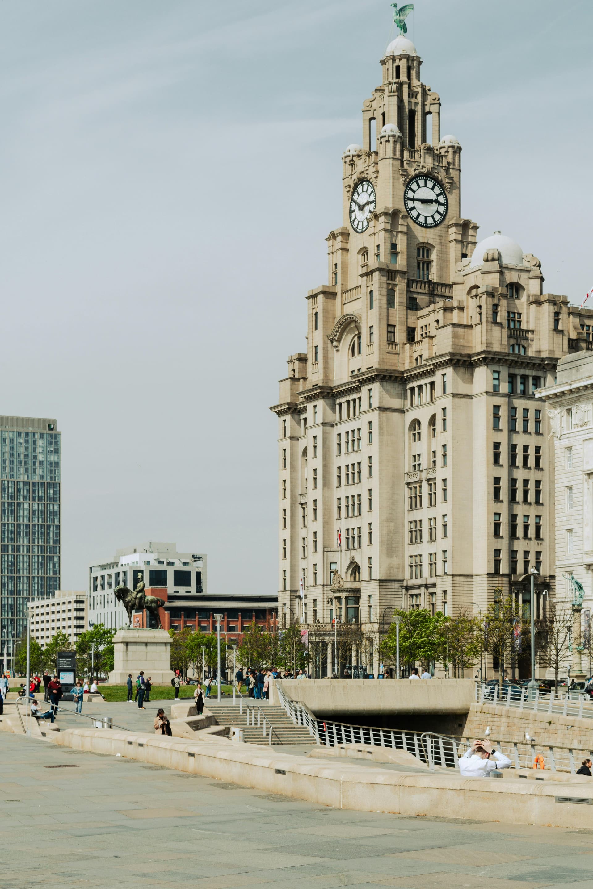 Historic clock tower building beside a paved walkway with people walking and sitting nearby.