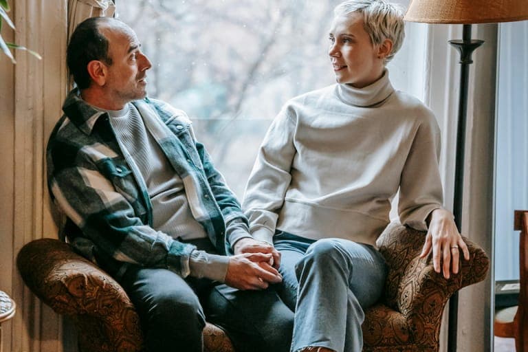 A man and a woman sit together on a sofa, holding hands and looking at each other, with a window in the background.