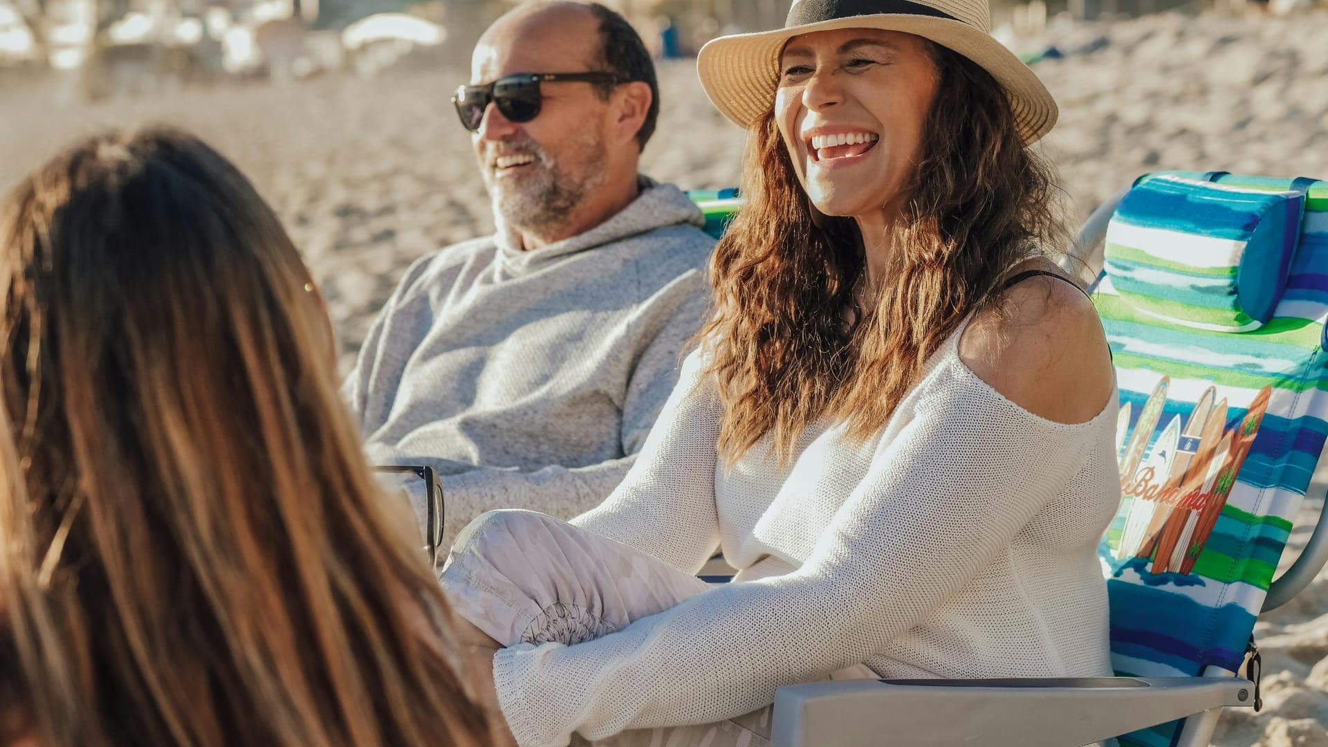 Two people sitting on a beach, smiling and enjoying the sun, with beach chairs and sand in the background.